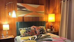 Un dormitorio con cabecera tapizada de verde, ropa de cama con estampado floral, un cuadro abstracto grande en la pared y un revestimiento de madera.