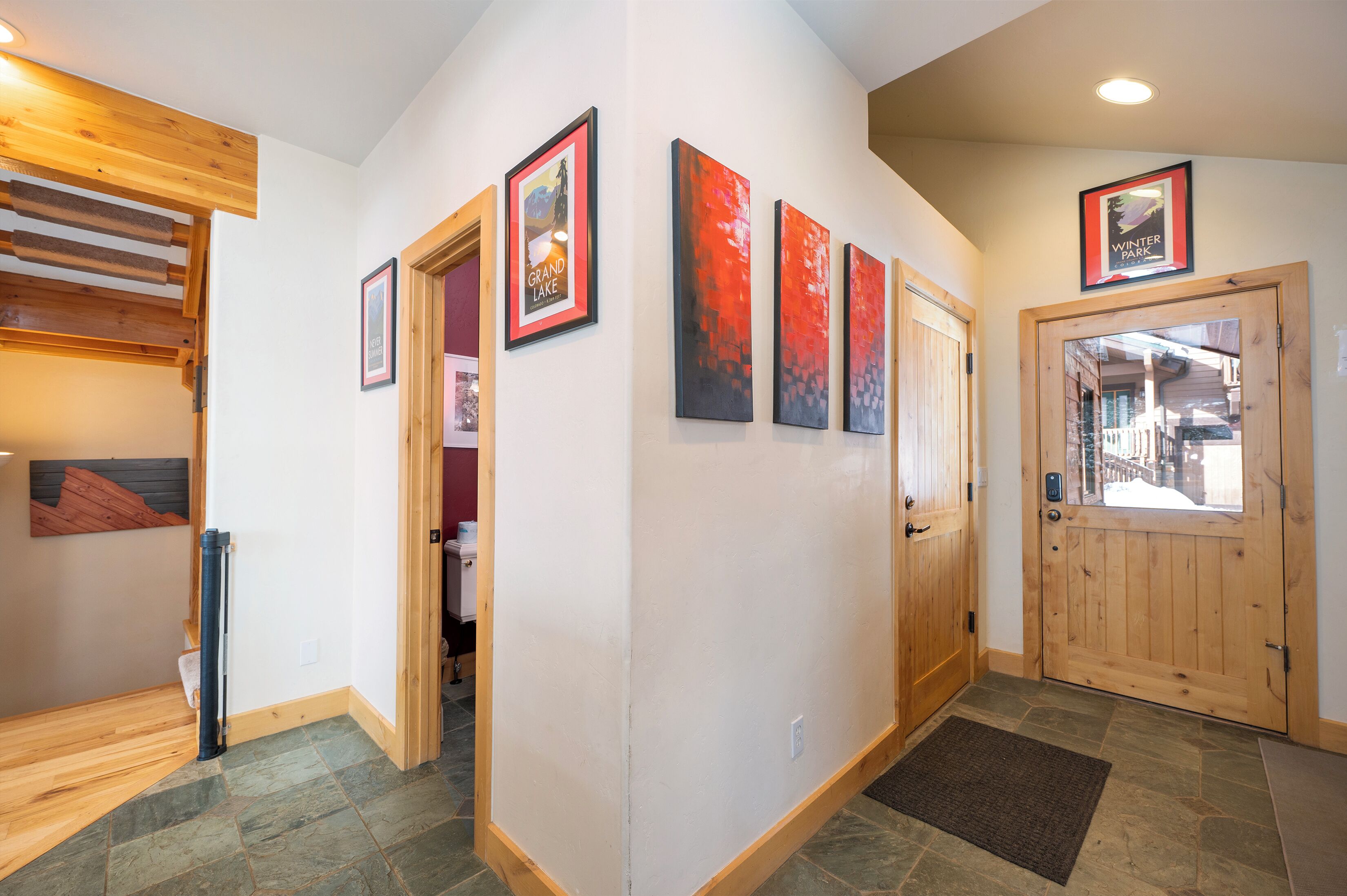 A hallway with wooden flooring, a wooden door, and a stone-tiled floor. There are framed pictures on the wall and a wooden beam structure on the left.
