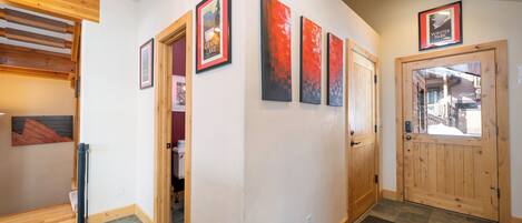 A hallway with wooden flooring, a wooden door, and a stone-tiled floor. There are framed pictures on the wall and a wooden beam structure on the left.