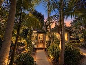 A house with a glass door entrance, surrounded by palm trees and a pathway leading to the door.