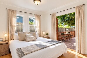 A bedroom with a large bed, two bedside lamps, a bedside table with a lamp, a window with curtains, and a view of an outdoor patio with a table and chairs. - Hammonds Beach Haven - next to Miramar Beach w/ yard, deck & sunroom - walk to Rosewood! (Santa Barbara)