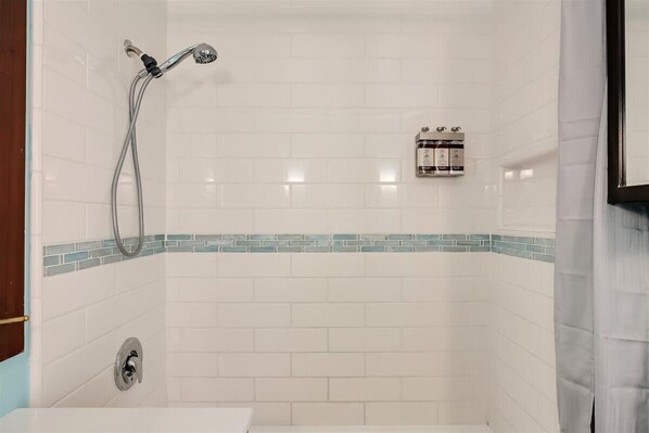 A bathroom with white tiled walls, a shower with a handheld showerhead, and a small bottle of shampoo on the wall.