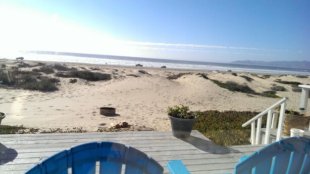 A beach view from a wooden deck with blue chairs, a fire pit, and a planter with plants.