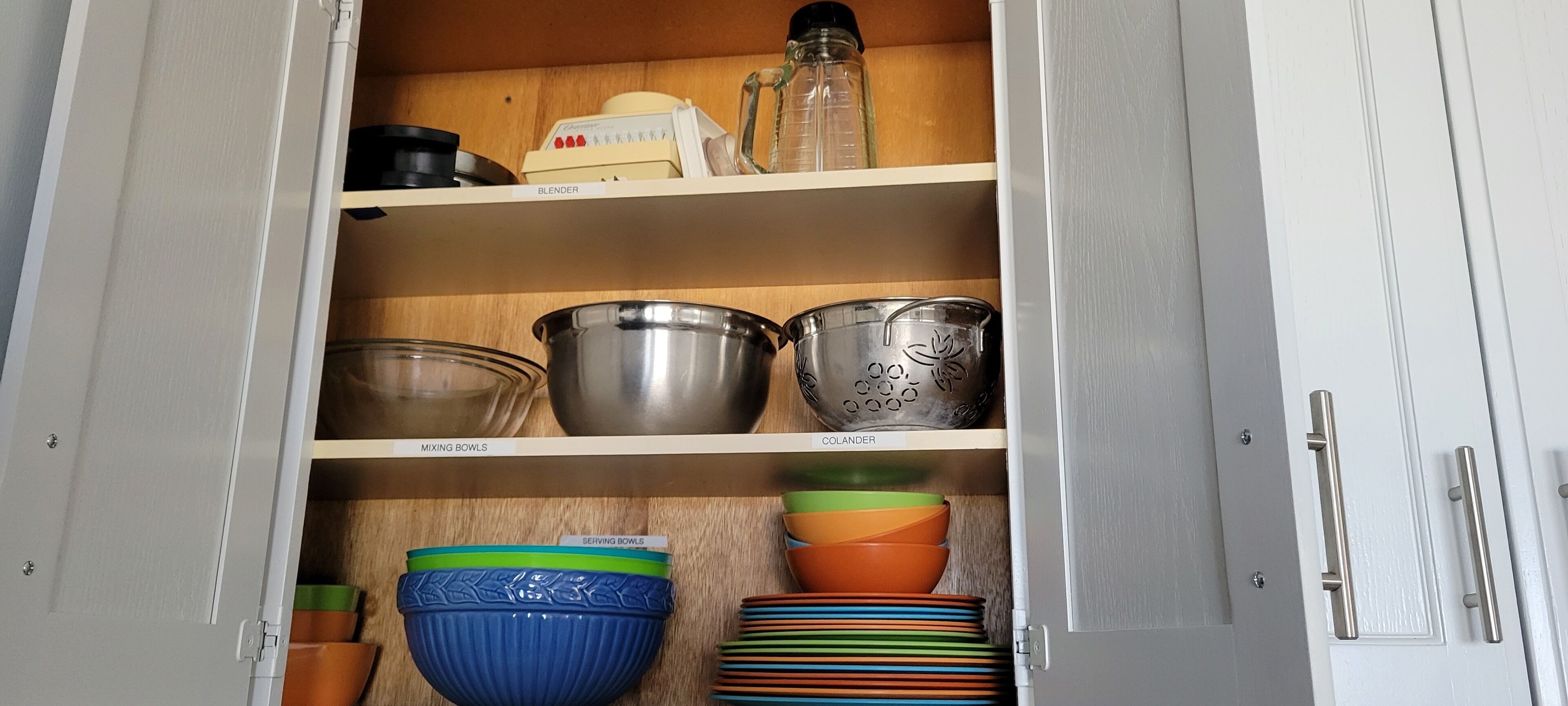 A kitchen cabinet with labeled shelves for mixing bowls, serving bowls, and colander.