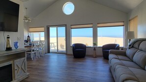 A spacious living area with a large TV, a wooden coffee table, and a beige sofa. There are two chairs and a small table with a lamp. The room has large windows with a view of the beach.