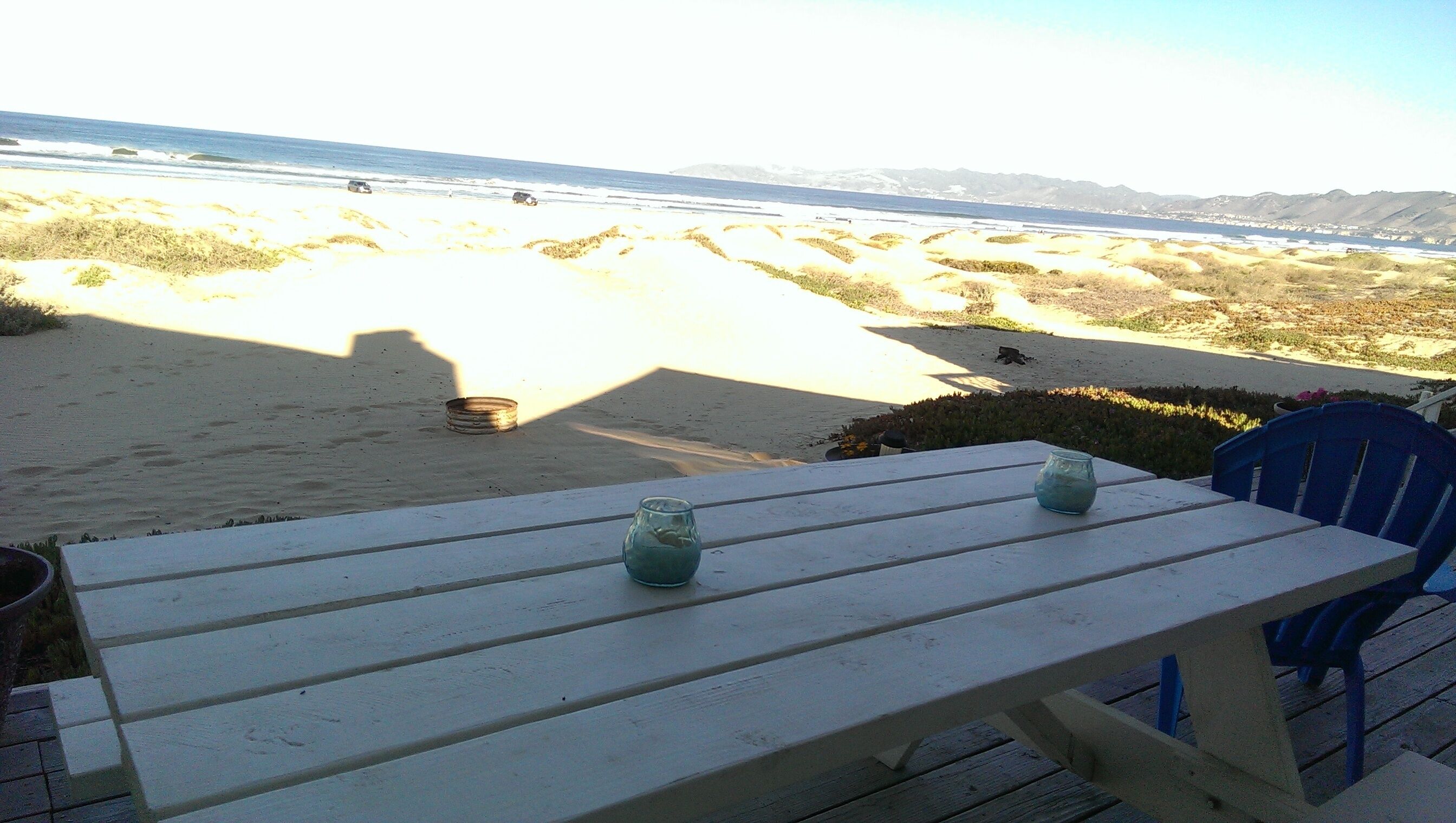 Ein Tisch direkt am Strand mit zwei Gläsern, mit Blick auf einen Sandstrand, Dünen und ferne Berge.