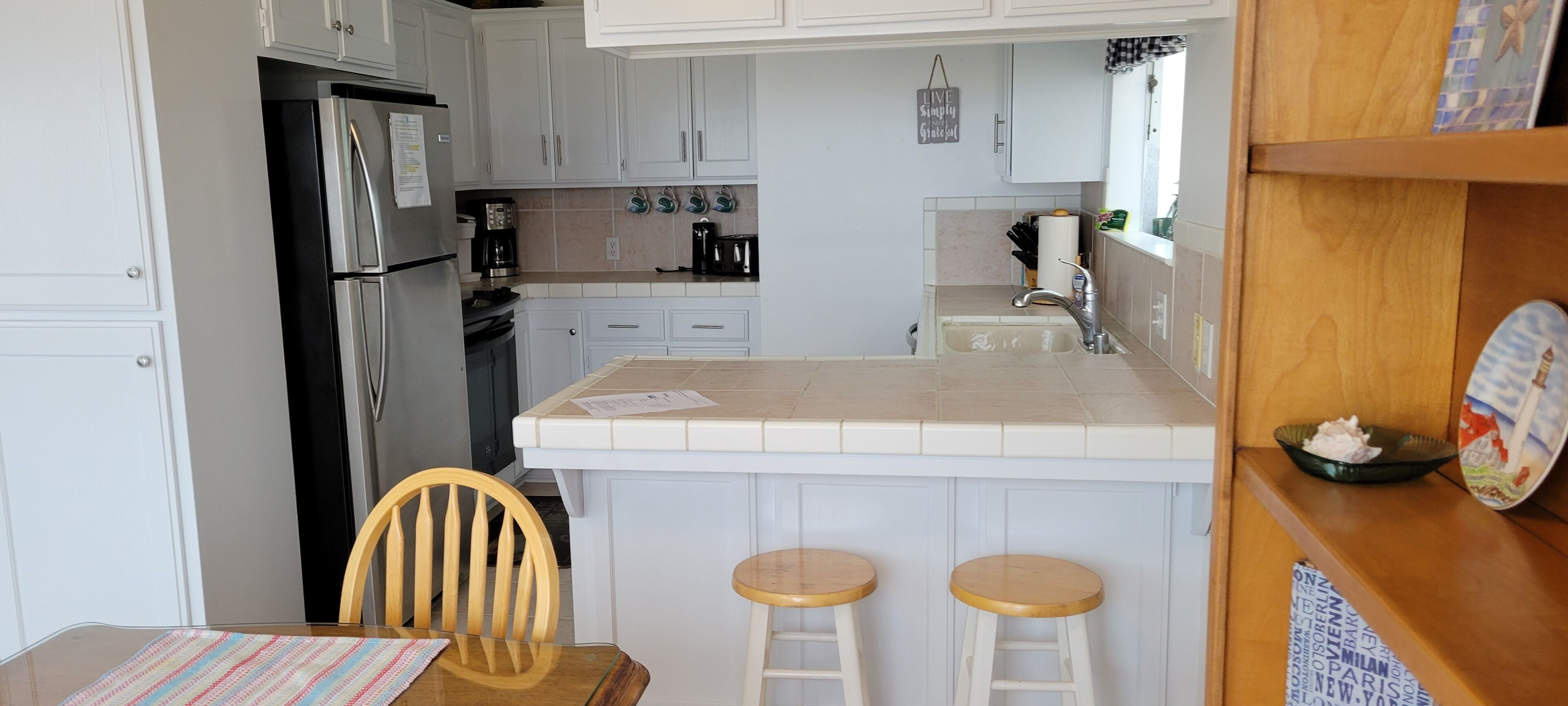 A compact kitchen with white cabinets, a stainless steel refrigerator, a coffee maker, and a sink. There are two stools and a wooden table with a striped tablecloth.
