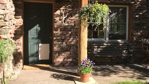 A rustic log cabin with a stone entrance, a wooden porch, and a hanging flower basket.