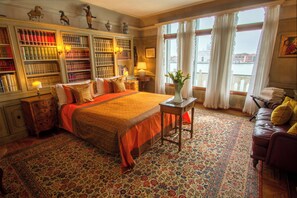A classic hotel room with a large bed, a bookshelf, a view of the city, and a leather armchair.