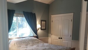 A bedroom with a wooden bed, a window with curtains, a door, and a wall-mounted lamp.