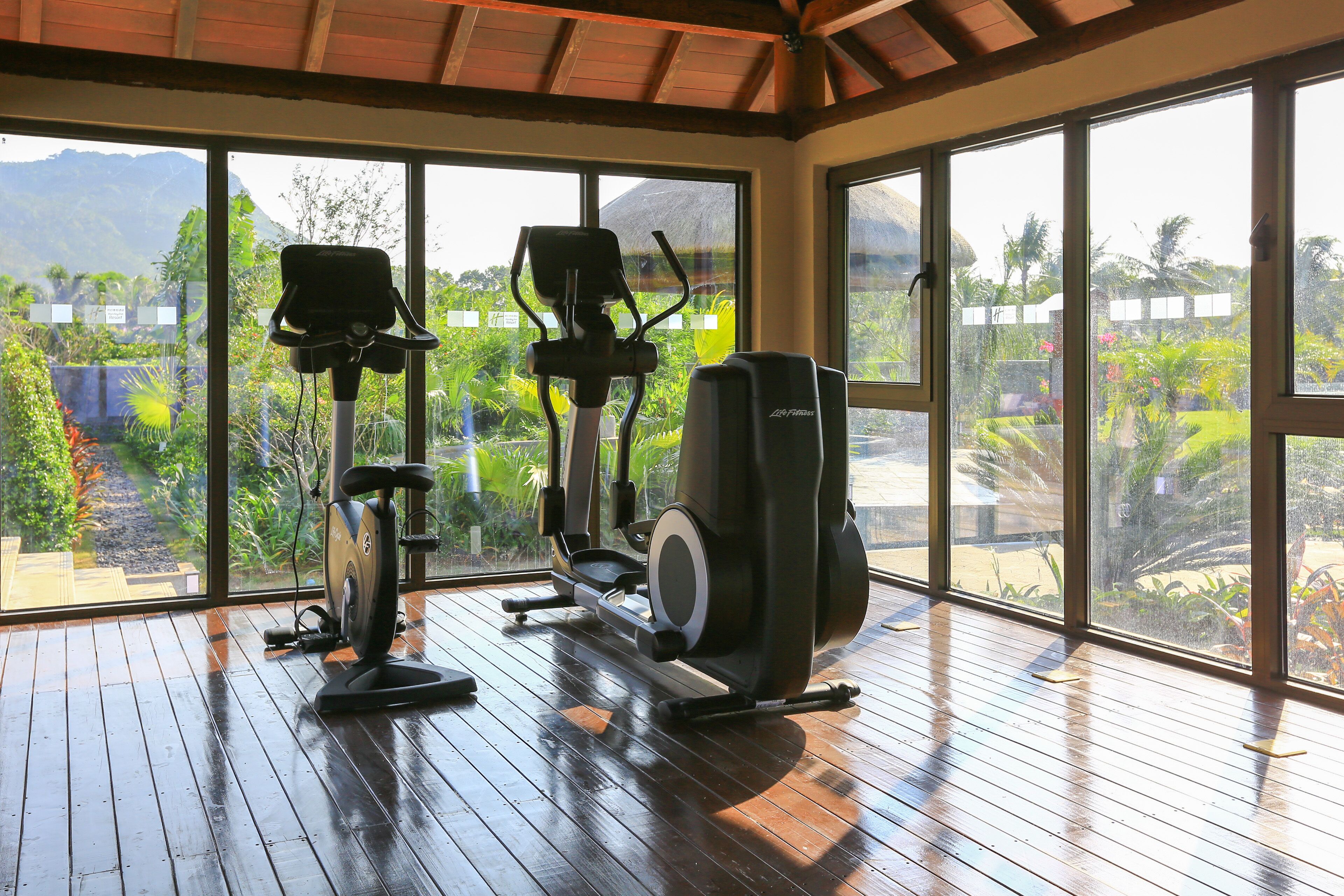 A gym with exercise equipment and large windows overlooking a garden.