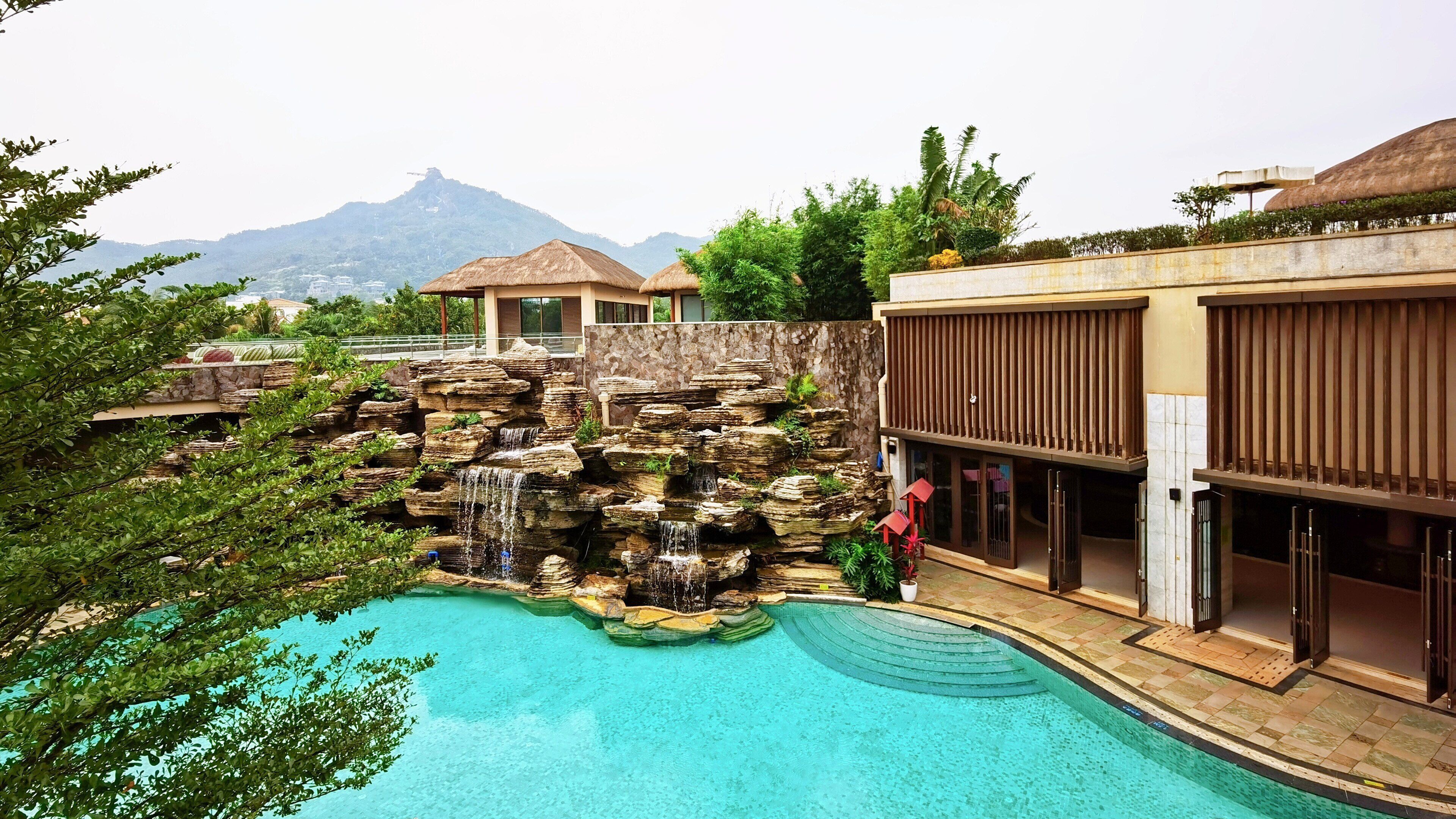 A swimming pool with a rock waterfall feature, surrounded by lush greenery and a mountain view.