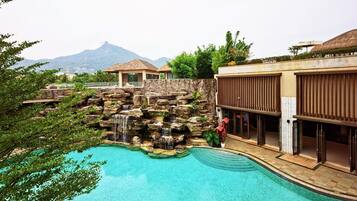 A swimming pool with a rock waterfall feature, surrounded by lush greenery and a mountain view.