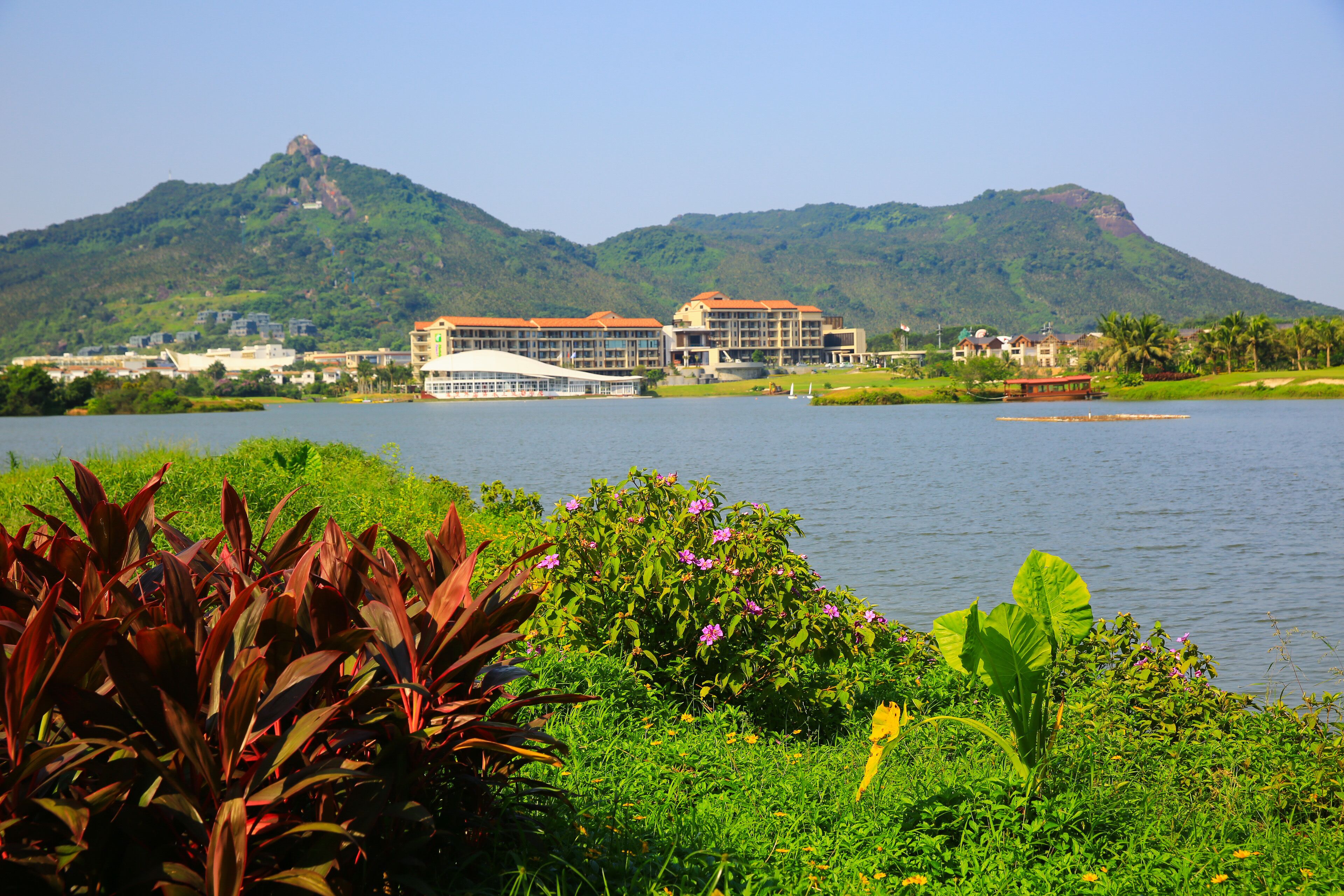 A hotel with a large building and a smaller structure across the water, surrounded by lush greenery and a mountain in the background.
