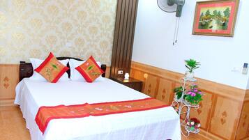 A neatly made bed with a white and red bedspread, two pillows, a bedside table with a lamp, a wall-mounted fan, and a framed picture on the wall.