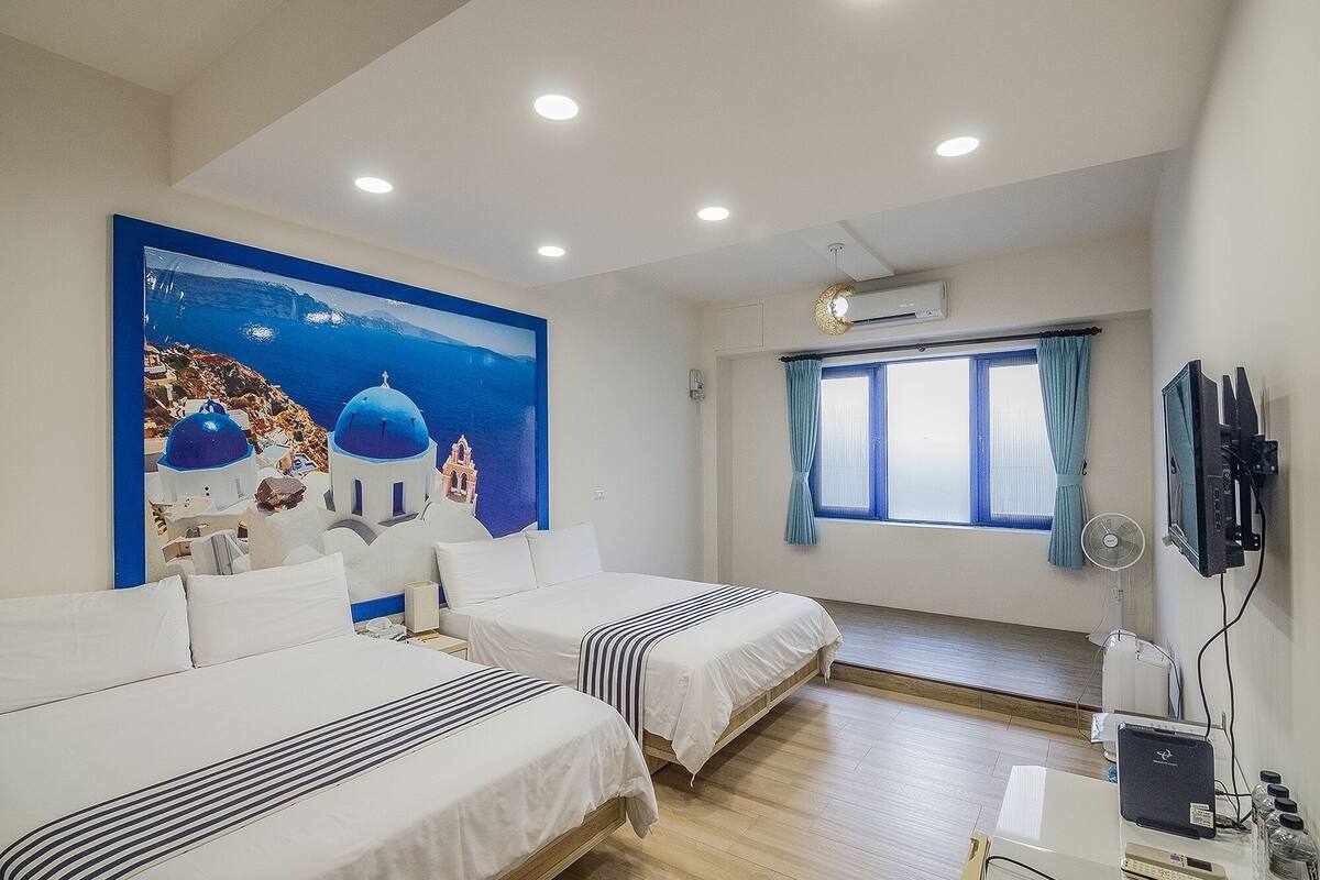 Een hotelkamer met twee bedden, een groot schilderij van een kustlandschap, een televisie en een raam met gordijnen.