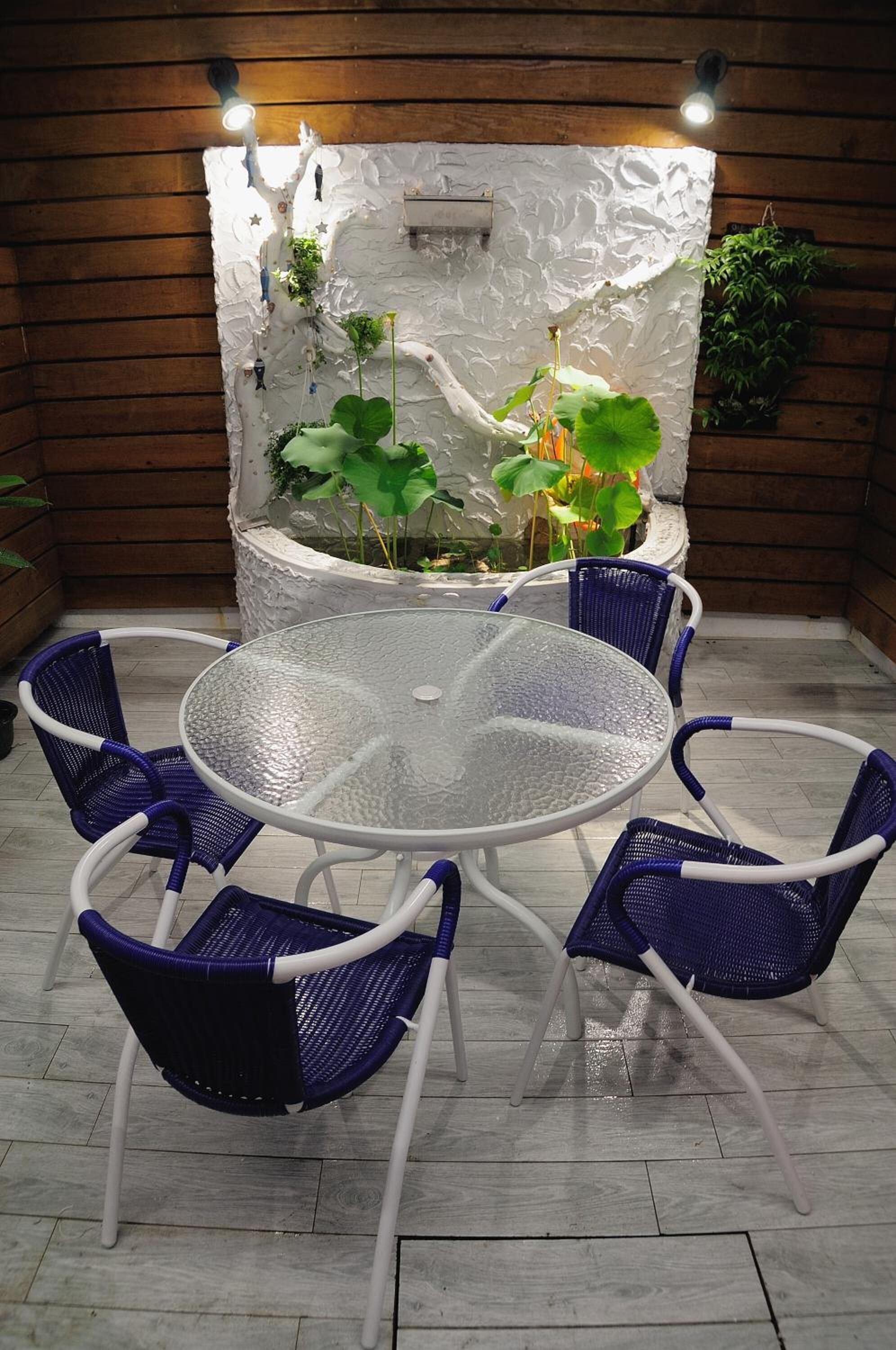 Une table ronde en verre, entourée de quatre chaises bleues, se trouve devant un mur blanc à texture, agrémenté de plantes, et le sol est en bois.