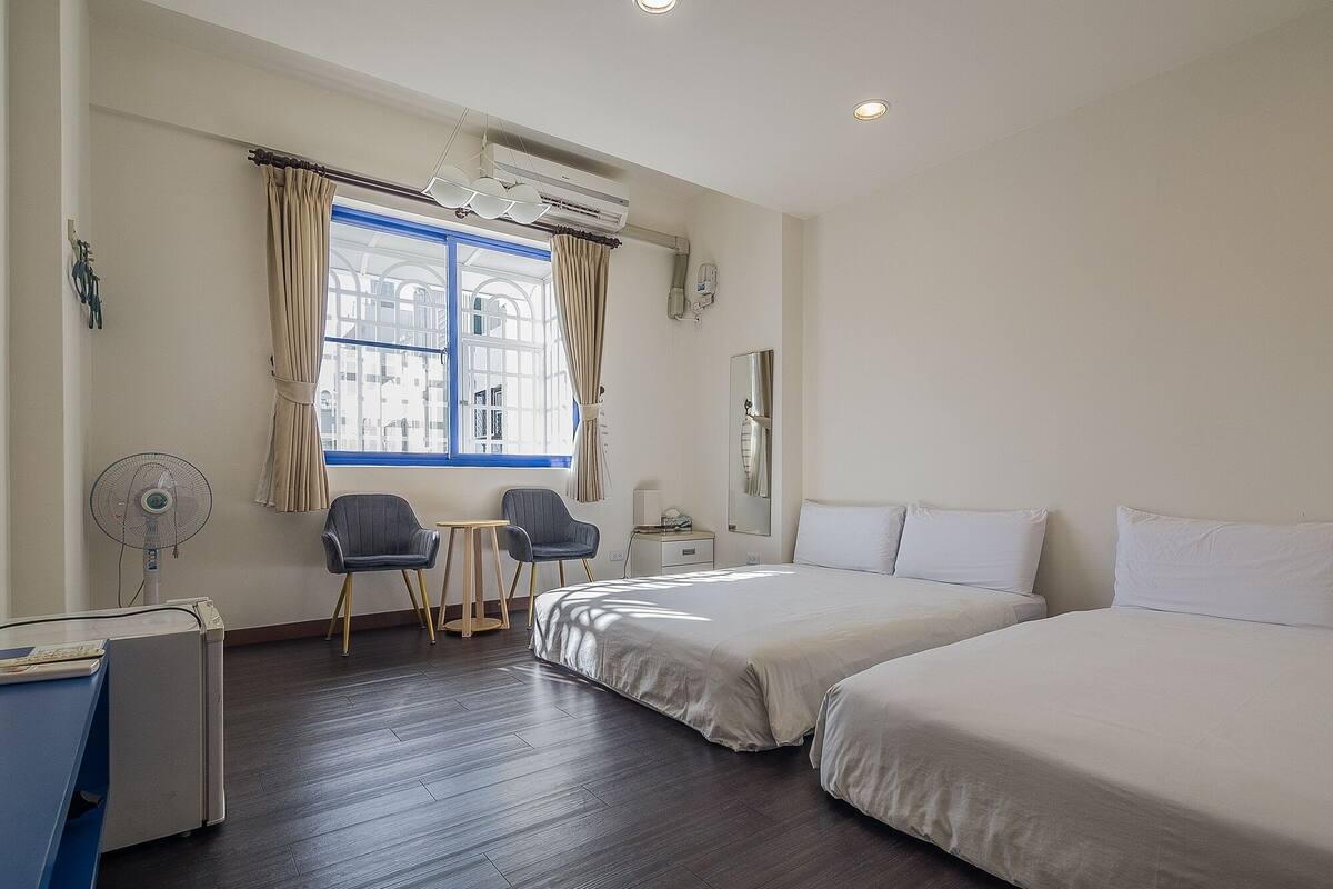Een hotelkamer met twee bedden, een bureau met stoel, een kleine tafel en een raam met gordijnen.