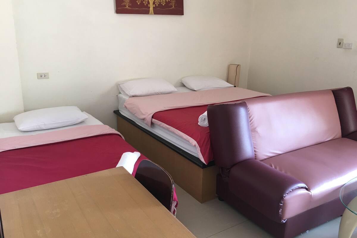 A hotel room with two beds, a wooden table, a leather sofa, and a wall-mounted power outlet.