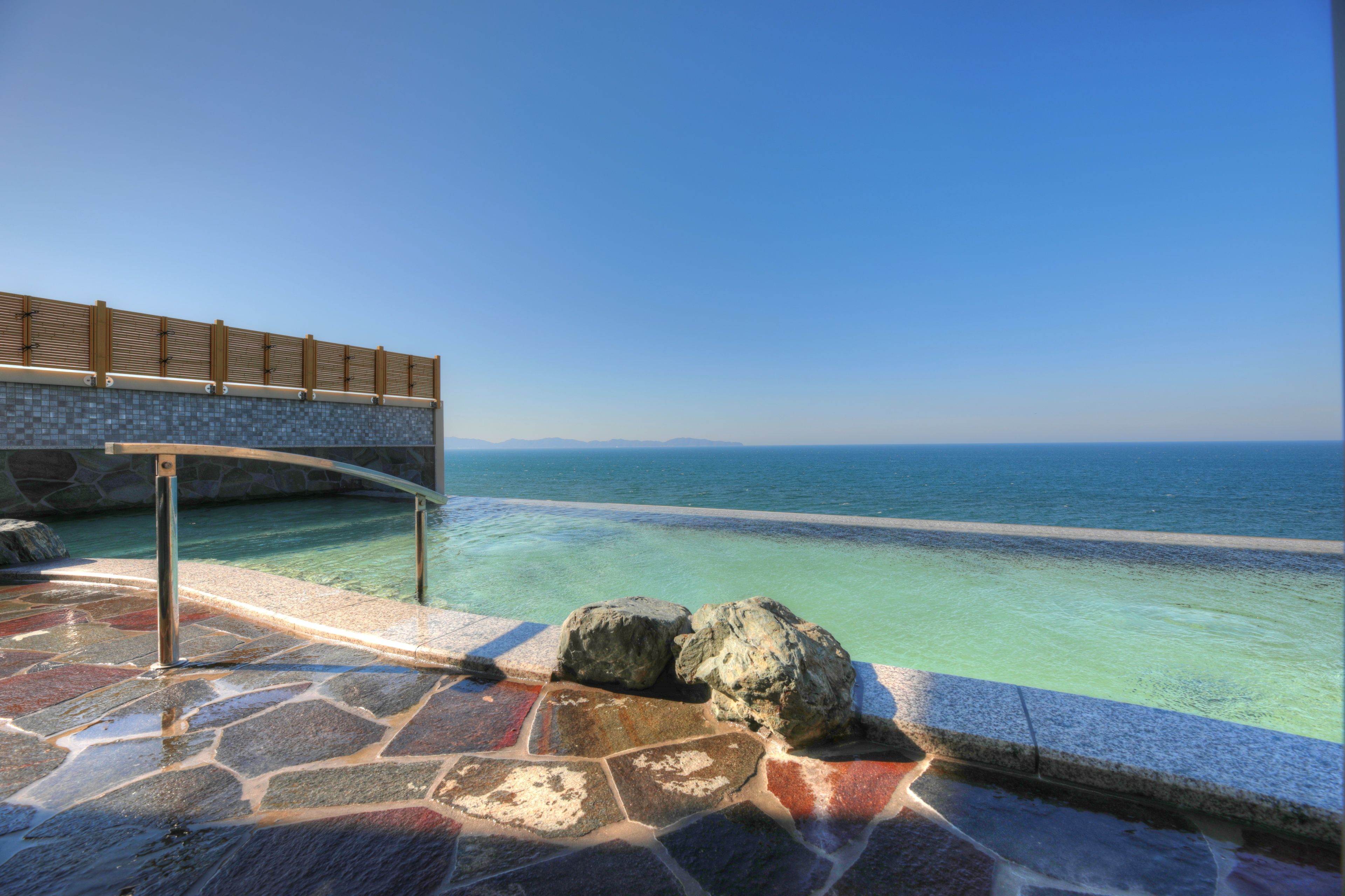 A stone pool with clear water, a wooden deck, and a view of the sea.