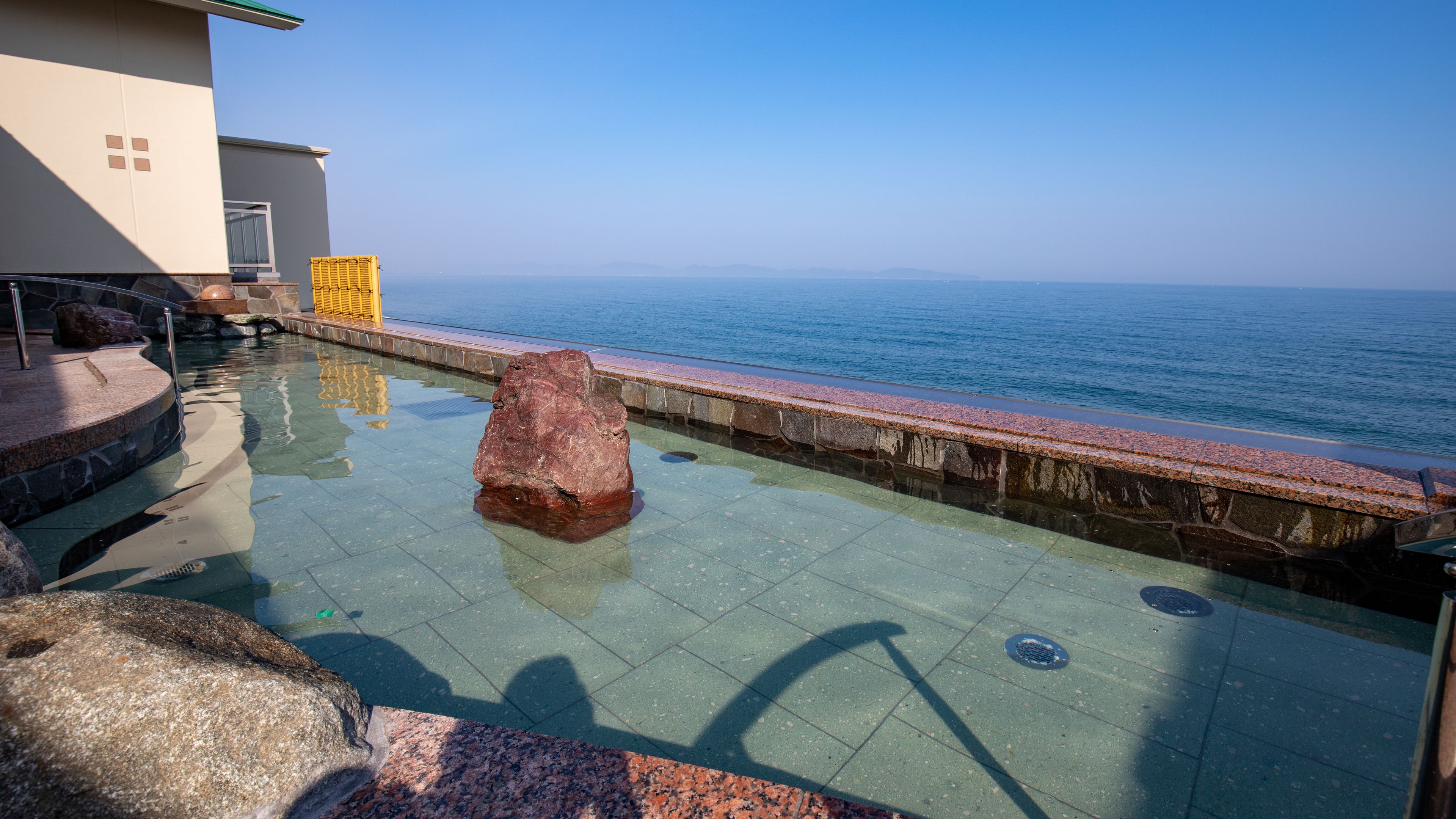 A pool area with a large rock centerpiece, a stone wall, and a view of the sea.
