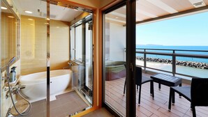 A modern bathroom with a glass-enclosed shower and a bathtub, leading to a balcony with a table and chairs overlooking a beach.
