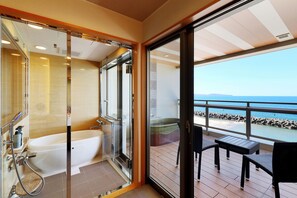 A modern bathroom with a glass-enclosed shower and a bathtub, leading to a balcony with a table and chairs overlooking a beach.