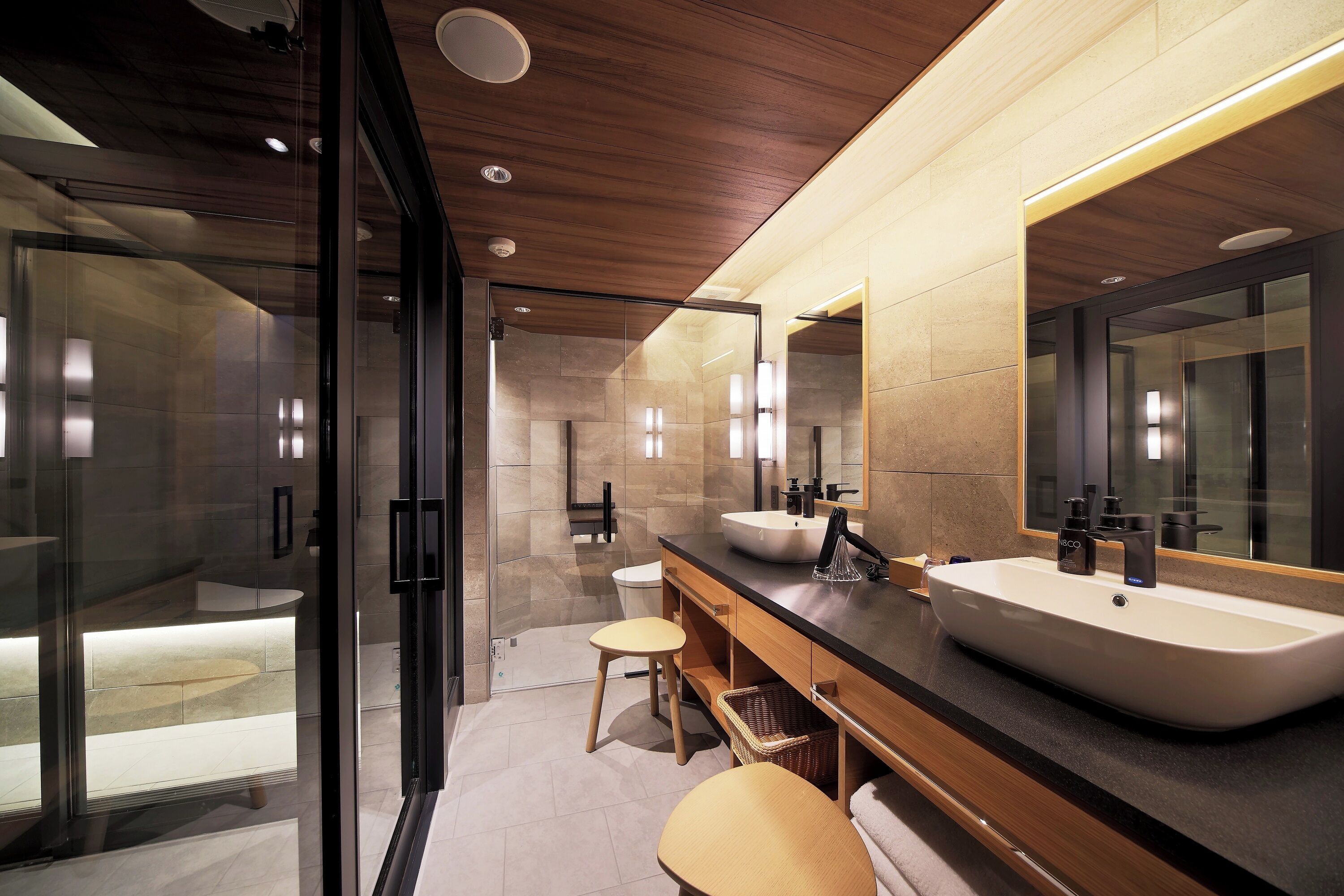 A modern bathroom with a large sink, two stools, a glass shower enclosure, and a wooden ceiling.