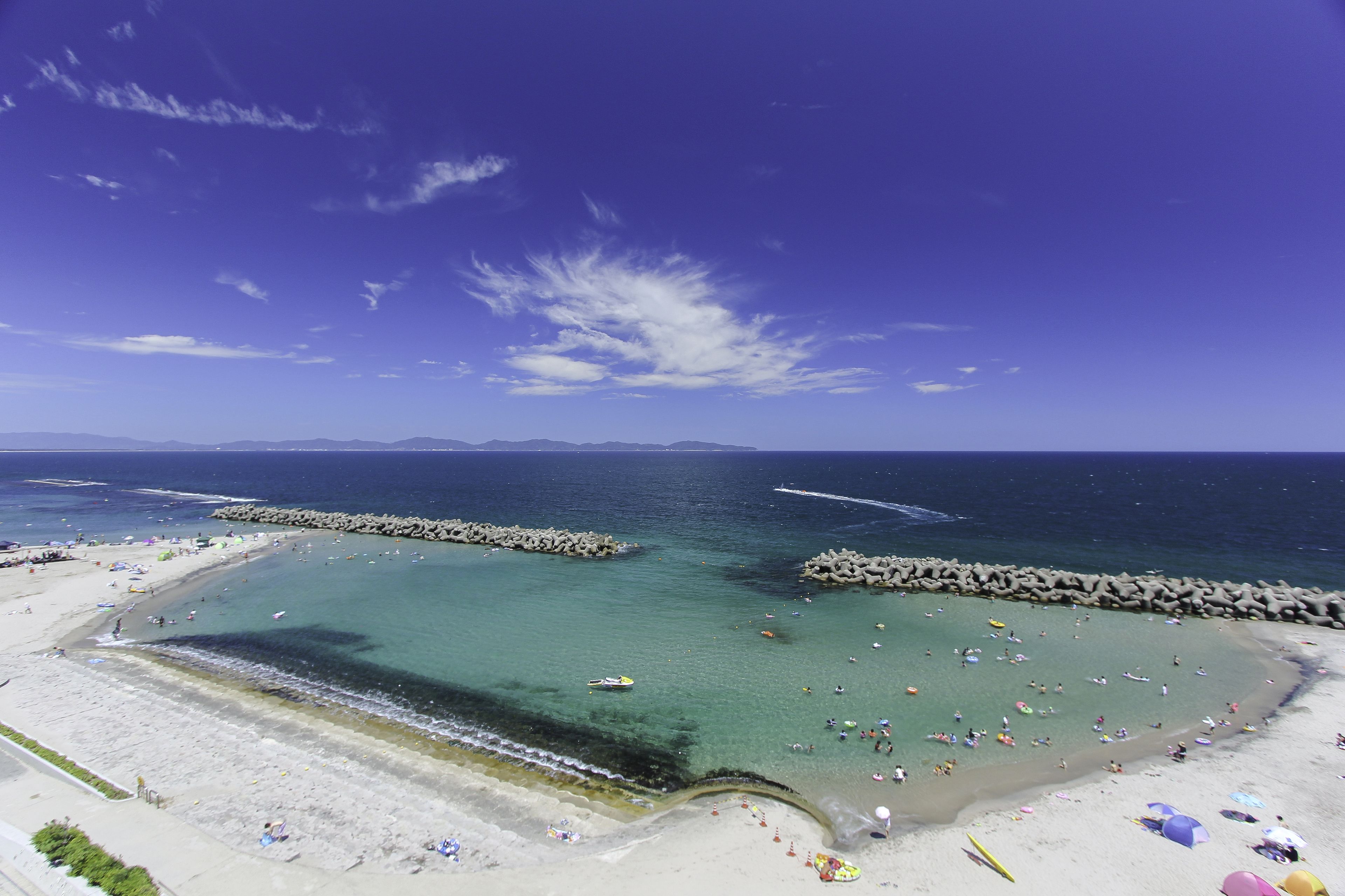 A beach with clear waters, rocky breakwaters, and a wide sandy area with umbrellas.