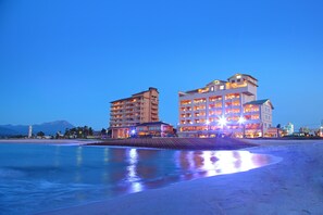 A beachfront hotel with multiple floors and balconies, a pool area, and a view of the ocean and mountains in the background.