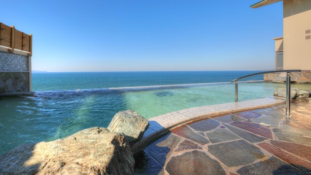 A stone pool with a clear view of the ocean, a stone deck, and a building with a flat roof.
