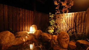 A nighttime garden with a wooden fence, rocks, a small pond, and a lit lantern.