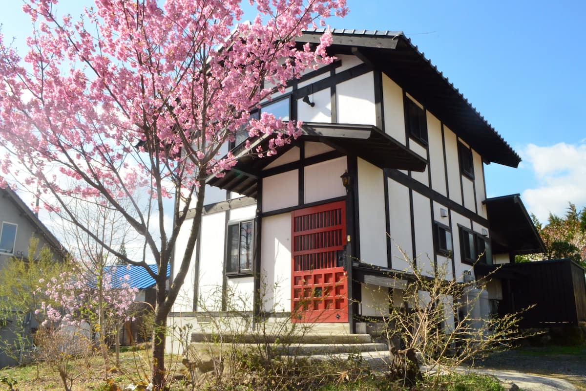 A traditional Japanese-style building with a red gate and a cherry blossom tree in front.