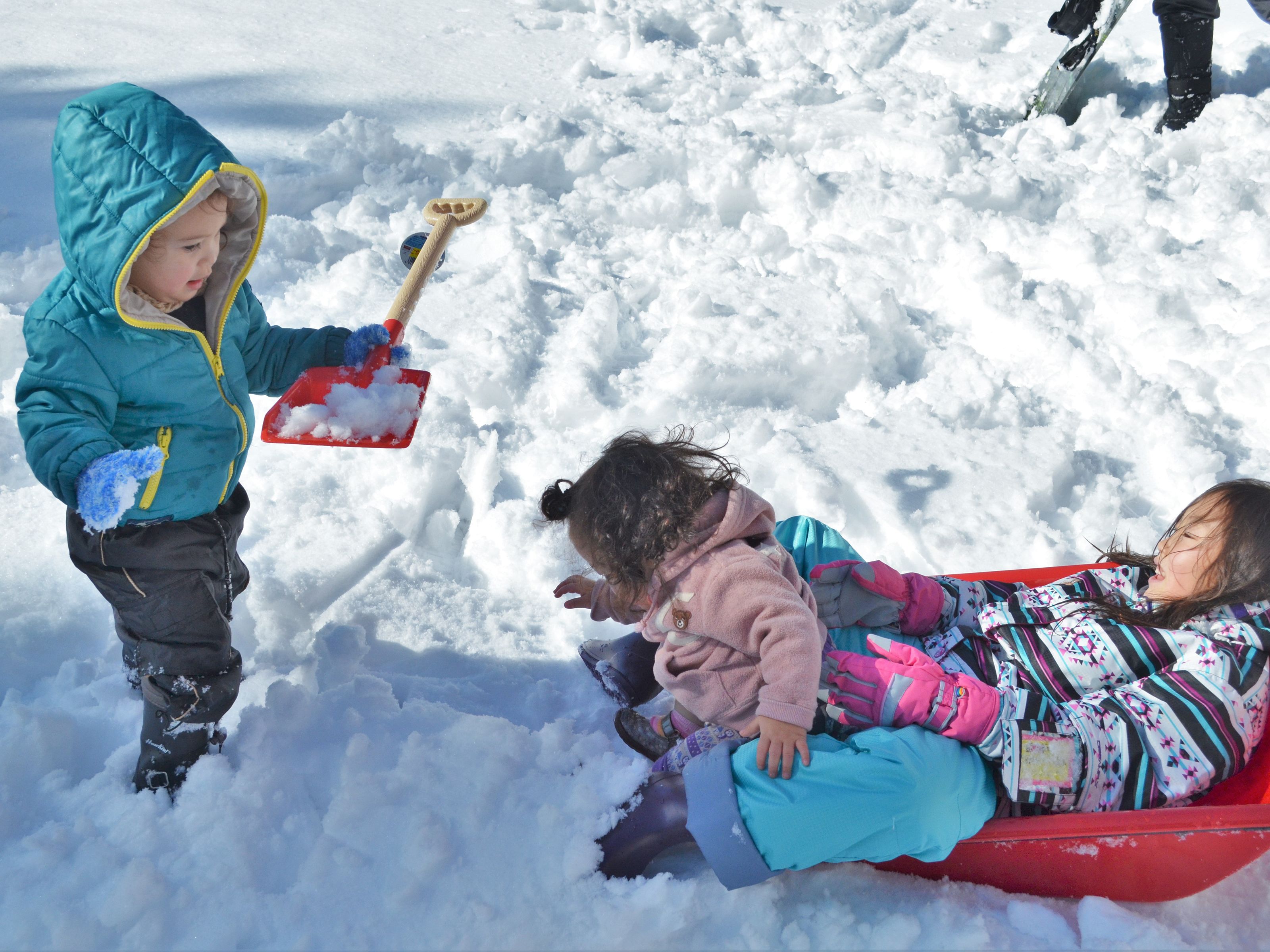 Two children playing in the snow, one standing and holding a shovel, and the other sitting on a sled.