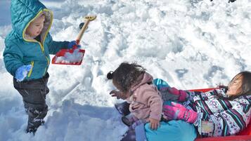 Two children playing in the snow, one standing and holding a shovel, and the other sitting on a sled.