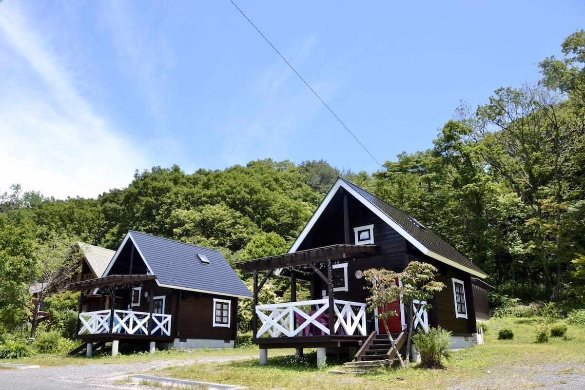 Two wooden cabins with white railings and a covered porch, surrounded by lush greenery.