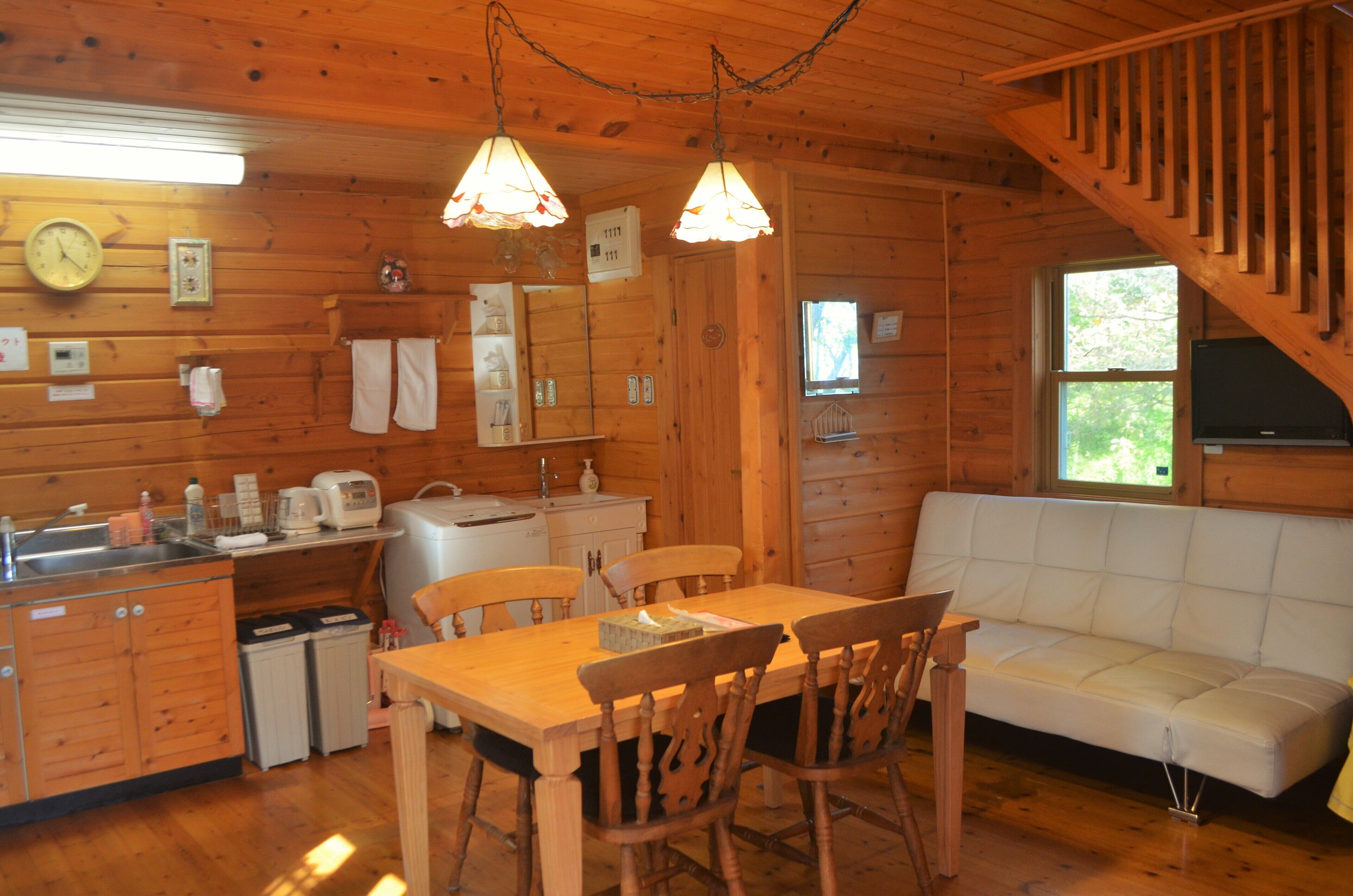 A wooden cabin interior with a dining table, chairs, a white sofa, a kitchen area with a sink, and a staircase.