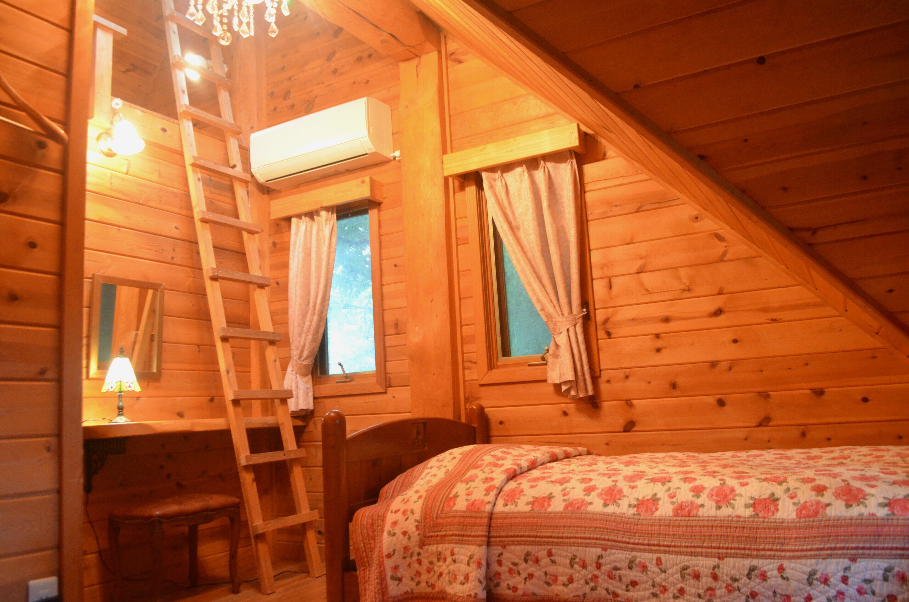 A wooden cabin room with a loft bed, a desk with a chair, a ladder, and an air conditioning unit.