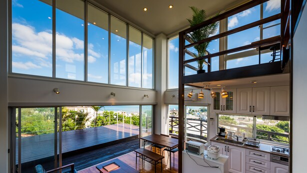 A spacious living area with a large window overlooking a balcony, a dining area, and a kitchen with white cabinets and a view of the city. - E-horizon Resort Condominium Yomitan (Yomitan)