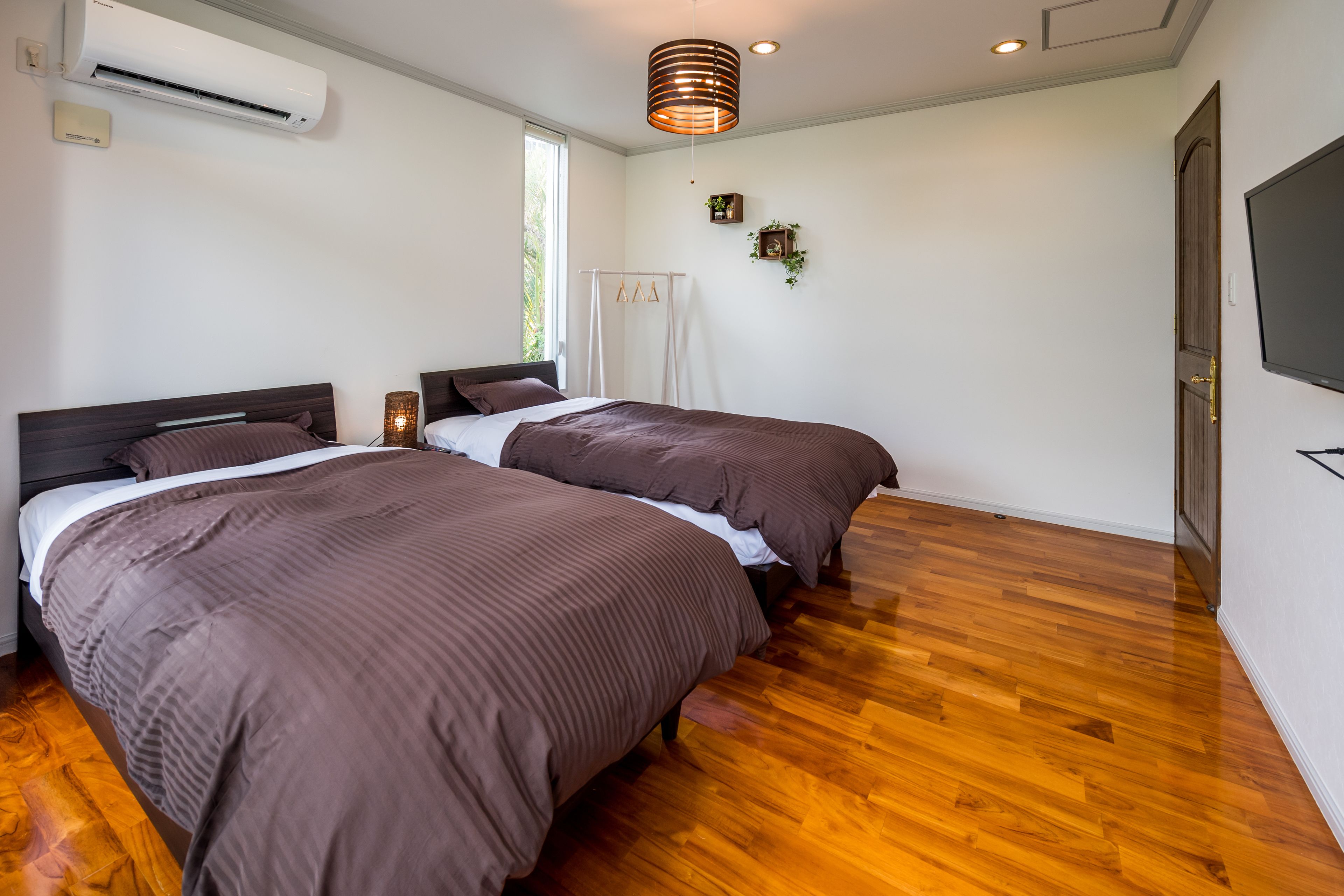 Un dormitorio con dos camas, suelo de madera, aire acondicionado, una lámpara de techo y televisor montado.