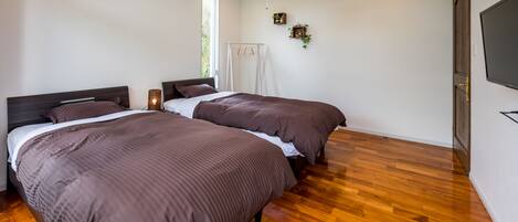 Un dormitorio con dos camas, suelo de madera, aire acondicionado, una lámpara de techo y televisor montado.
