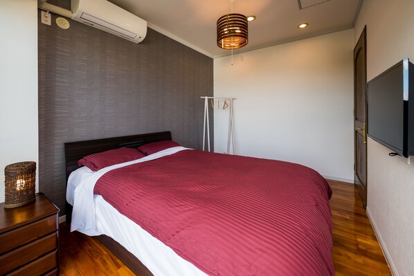 A modern bedroom with a large bed, a wooden dresser, a flat-screen TV, and a ceiling-mounted air conditioner.