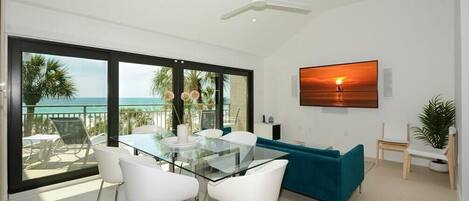 A modern dining area with a glass table and white chairs, a teal sofa, and a large window with a view of the ocean.