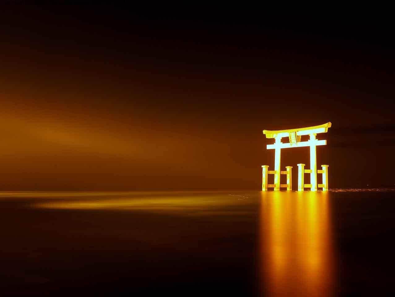 A traditional torii gate structure illuminated against a dark sky, with a reflective water surface in the foreground.