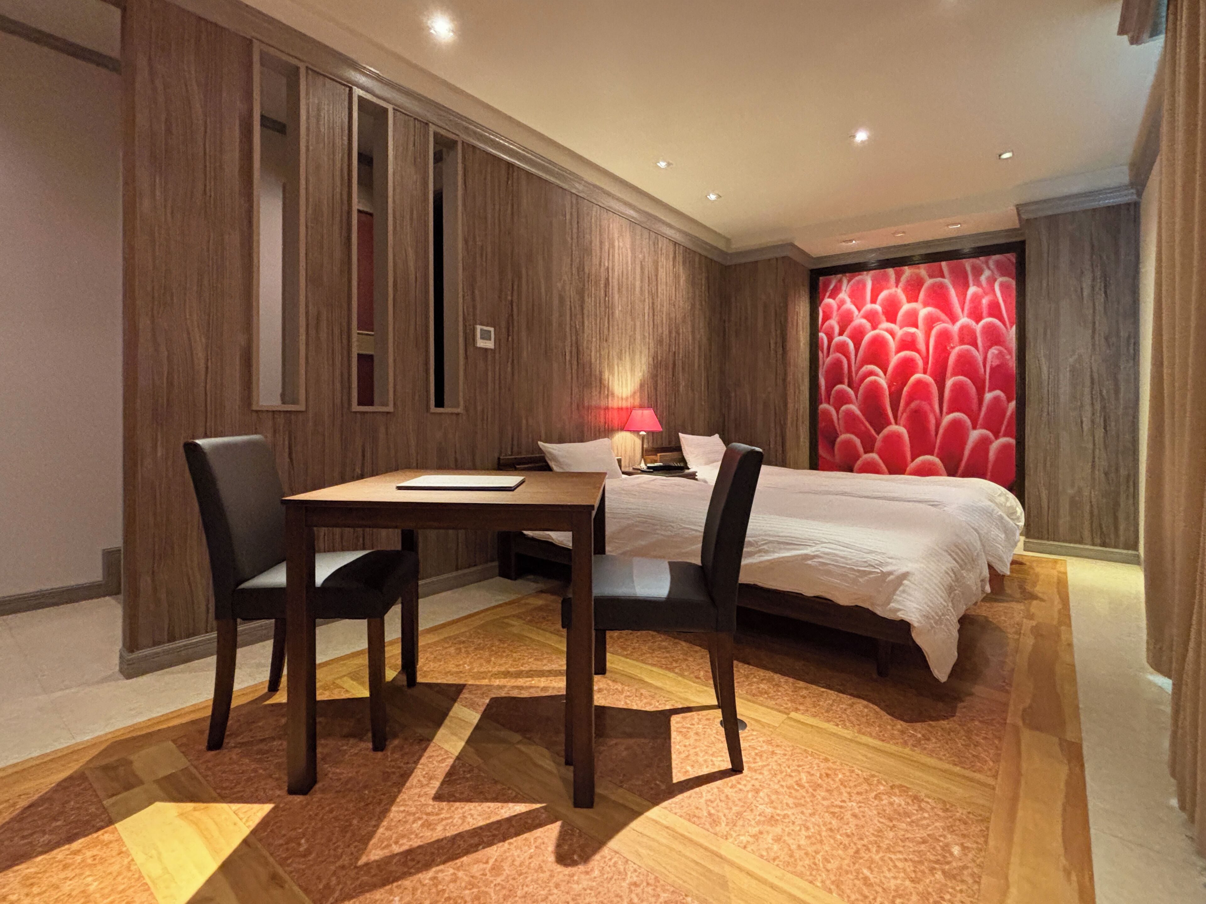 A hotel room with a bed, a desk, and two chairs. There is a red floral wallpaper on one wall and wooden paneling on the other.