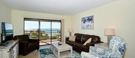 A living room with a large sliding glass door leading to a balcony, a brown leather sofa, a floral armchair, and a coffee table.