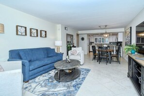 A living room with a blue sofa, a white armchair, a round coffee table, and a dining area with a wooden table and chairs.