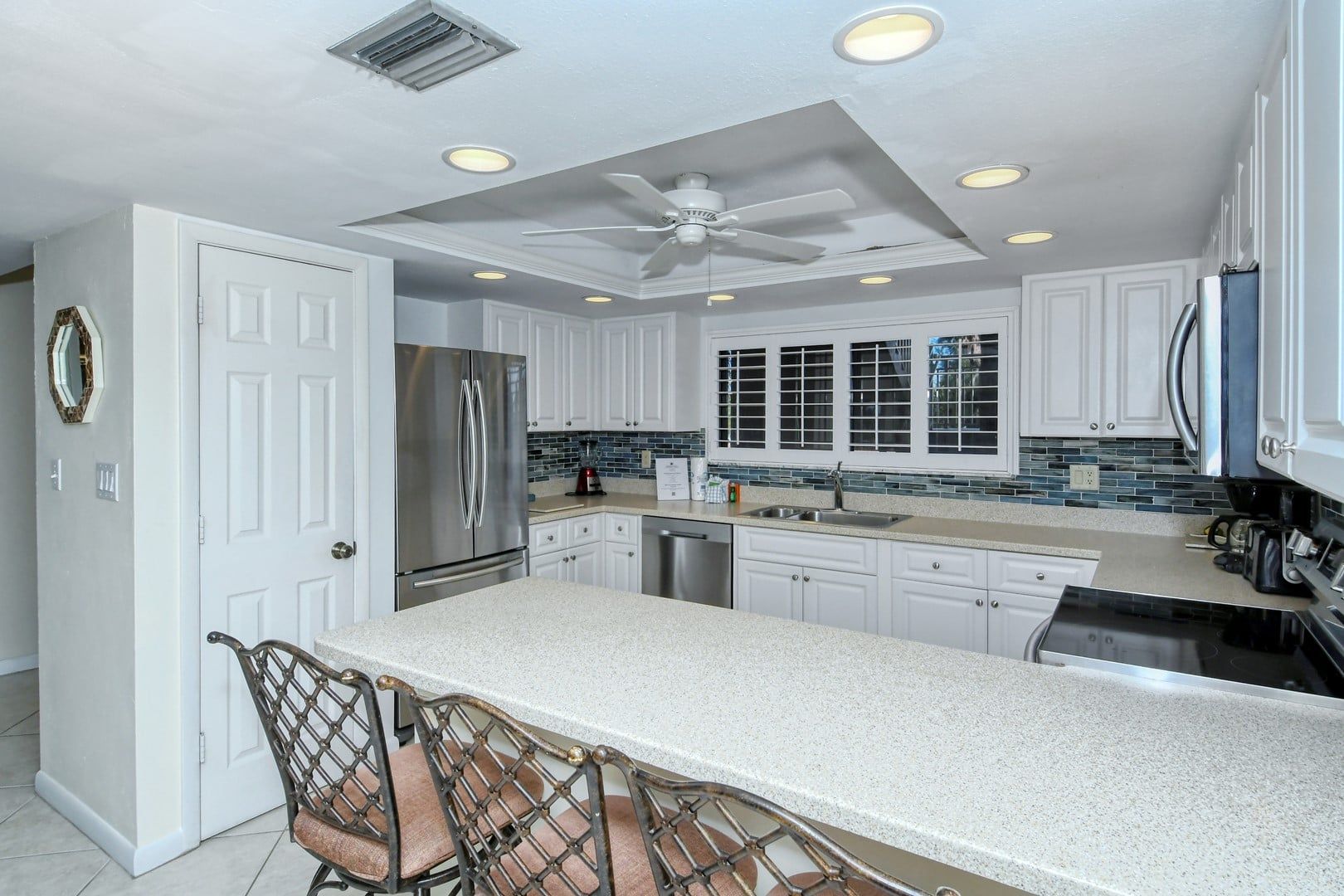 A modern kitchen with white cabinetry, a stainless steel refrigerator, a built-in oven, a dishwasher, and a bar area with a seating arrangement.