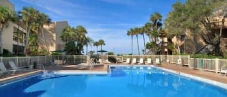 A swimming pool with lounge chairs and palm trees, surrounded by buildings.