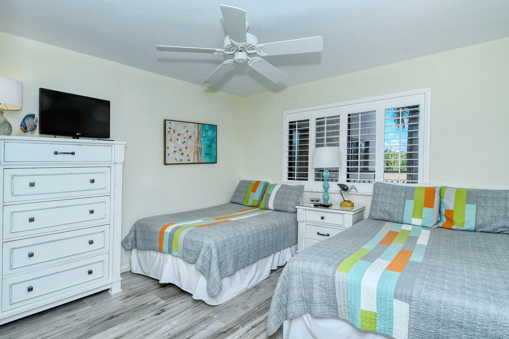A bedroom with two beds, a ceiling fan, a TV, a dresser, and a window with blinds.